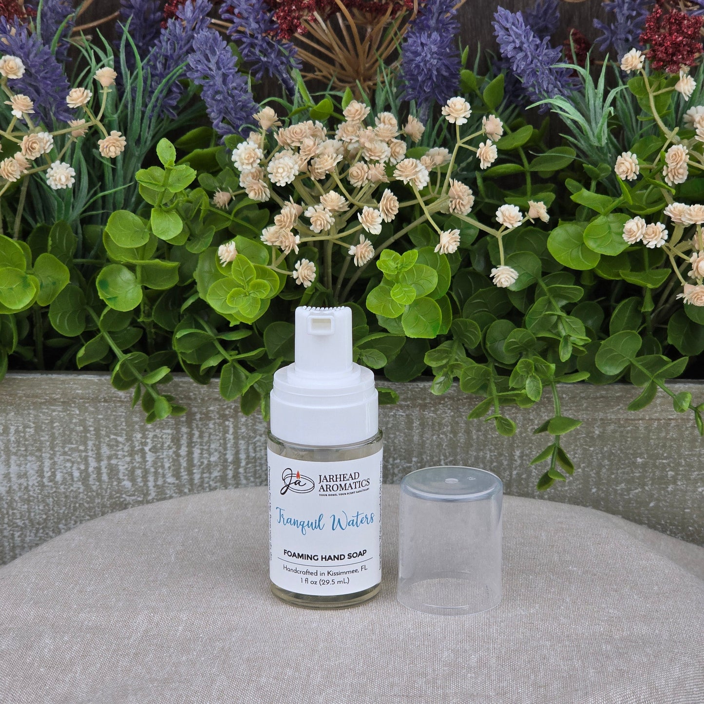 A bottle of Jarhead Aromatics Tranquil Waters Foaming Hand Soap, cap off, rests on a table before green leaves and purple-white flowers.