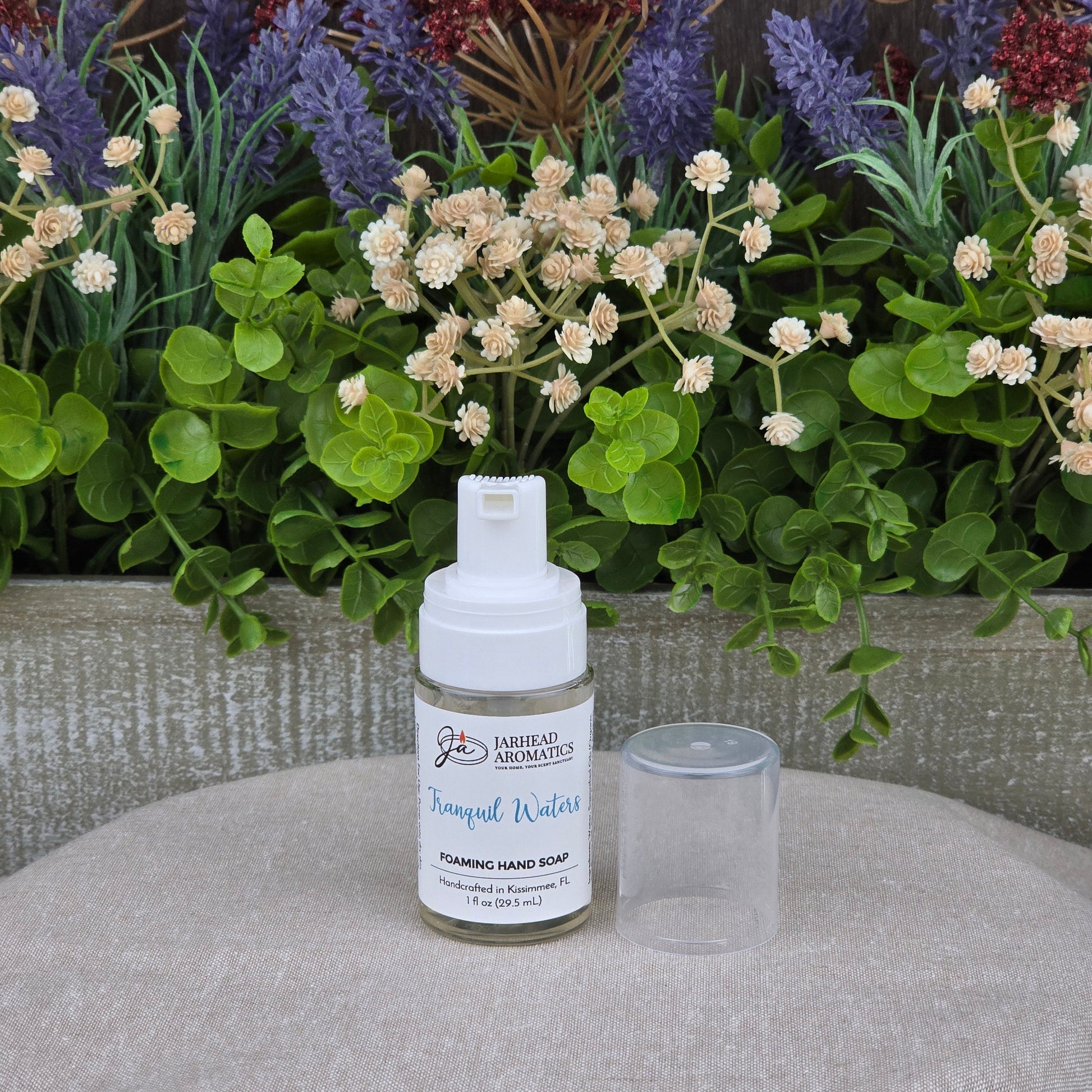 A bottle of Jarhead Aromatics Tranquil Waters Foaming Hand Soap, cap off, rests on a table before green leaves and purple-white flowers.