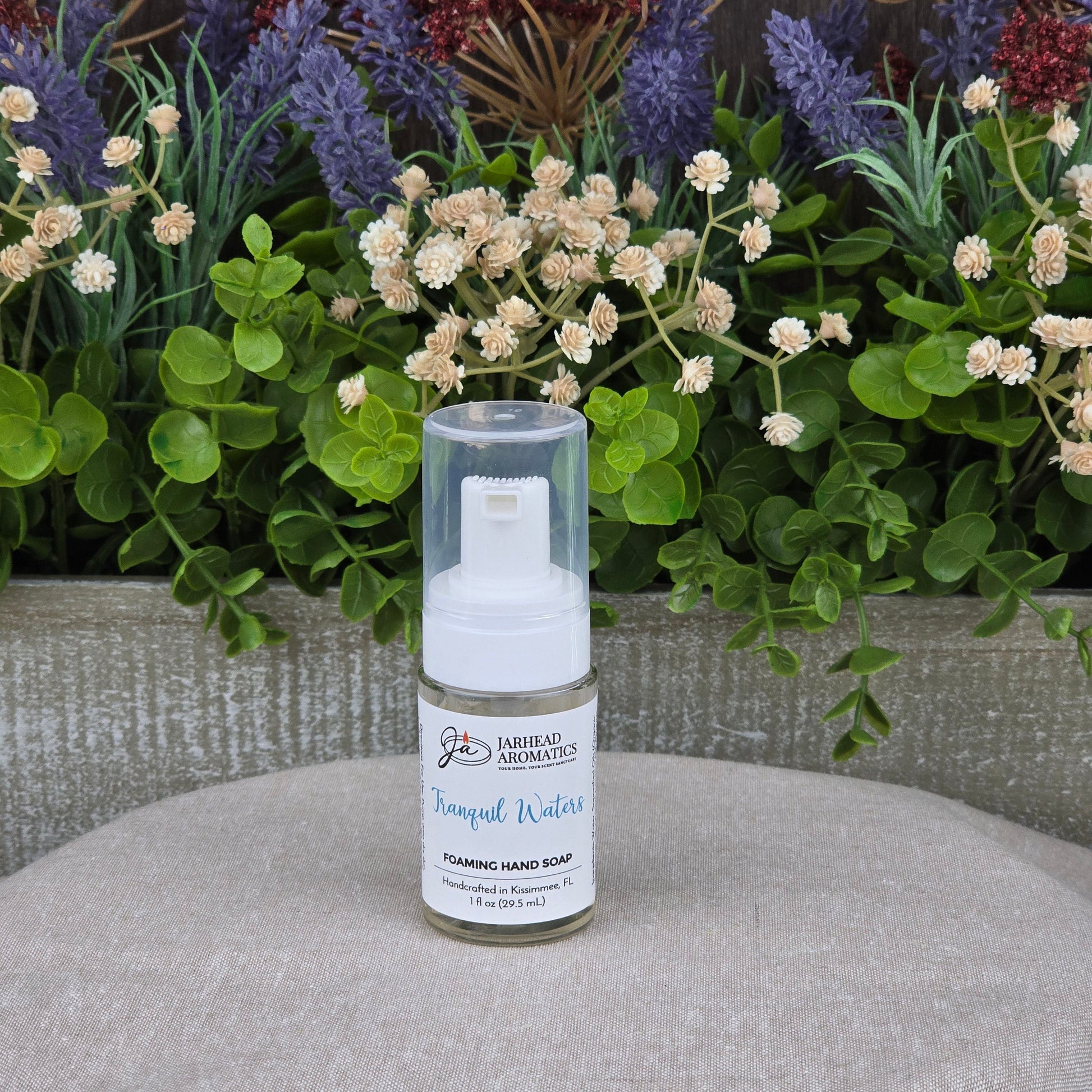 A bottle of Jarhead Aromatics Tranquil Waters Foaming Hand Soap rests on fabric, with green foliage and colorful artificial flowers in the background.
