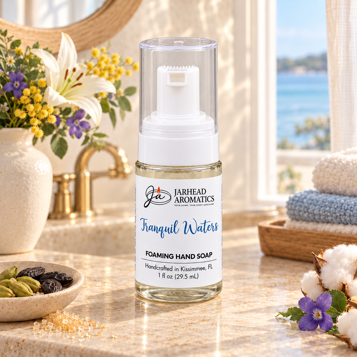 A bottle of Tranquil Waters Handmade Foaming Hand Soap with a clear pump dispenser, centered on a bathroom vanity beside a small bowl of spices, a stack of towels, and a vase of flowers.