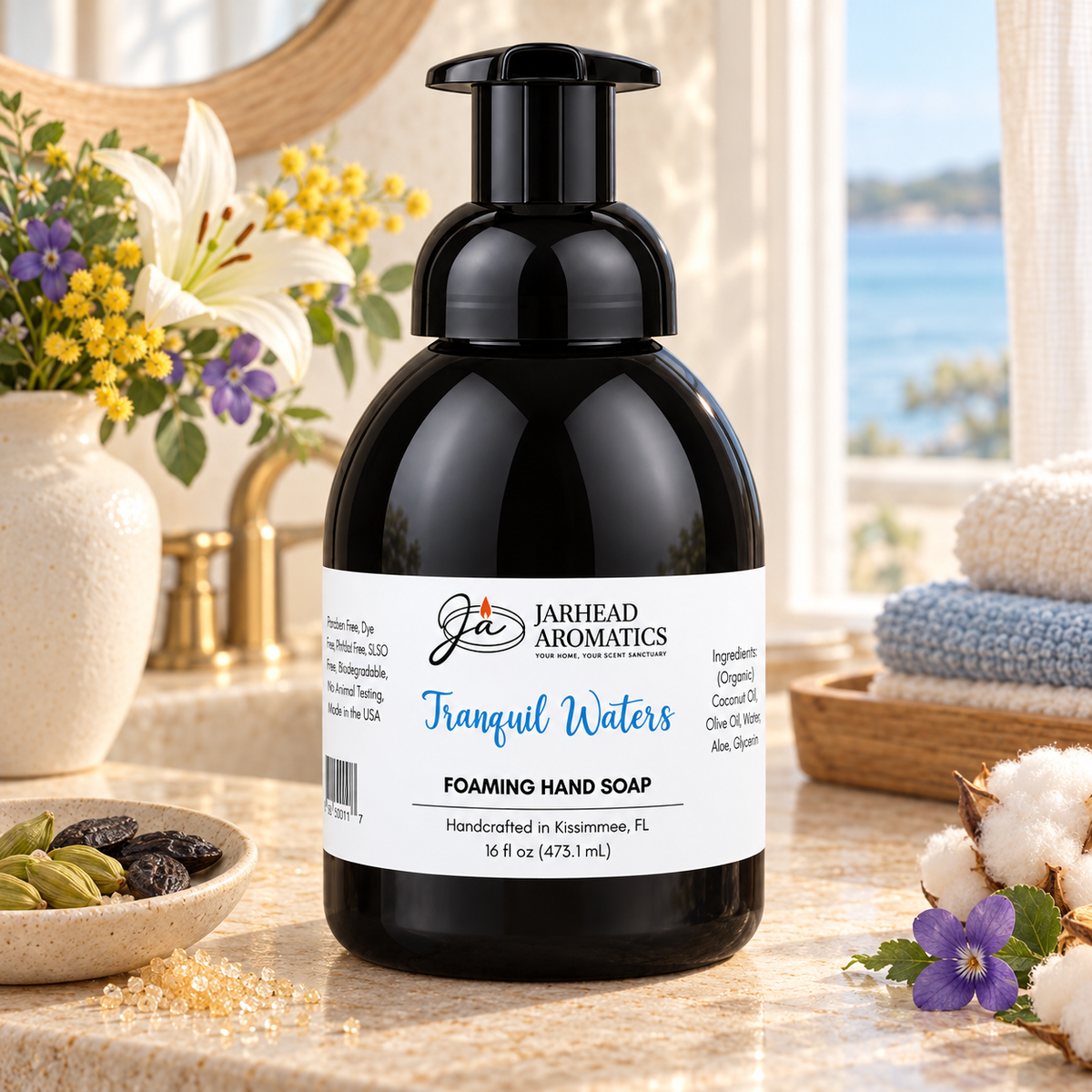 A black bottle of Tranquil Waters Handmade Foaming Hand Soap | Marine, Lily & Tonka Bean by Jarhead Aromatics sits on a bathroom counter near flowers and stacked towels, with a label displaying product name, ingredients, and company information.