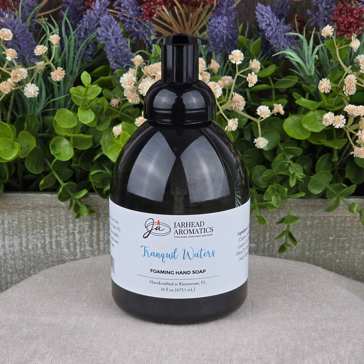 A bottle of Jarhead Aromatics Tranquil Waters Foaming Hand Soap rests on a textured surface, framed by green plants and purple flowers in the background.