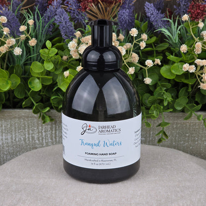 A bottle of Jarhead Aromatics Tranquil Waters Foaming Hand Soap rests on a textured surface, framed by green plants and purple flowers in the background.