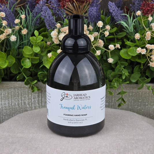 A bottle of Jarhead Aromatics Tranquil Waters Foaming Hand Soap rests on a textured surface, framed by green plants and purple flowers in the background.