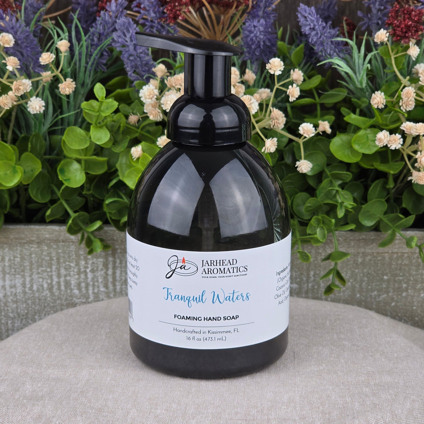 A black pump bottle of Jarhead Aromatics Tranquil Waters Foaming Hand Soap sits on a beige surface with green plants and purple and white flowers behind it. The label highlights its handcrafted quality in Macclenny, FL.