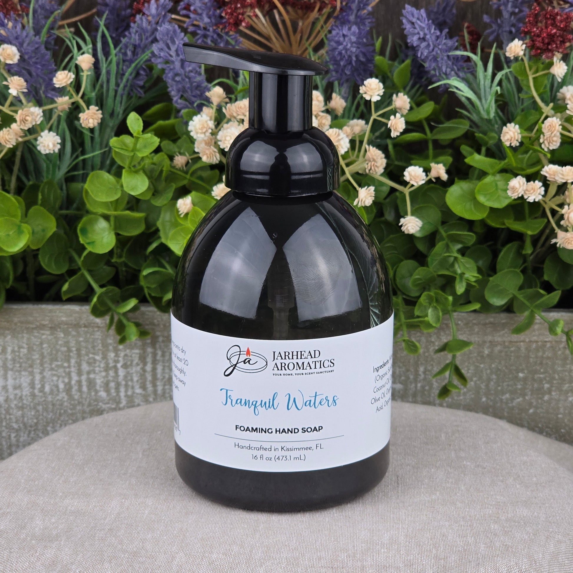 A black pump bottle of Jarhead Aromatics Tranquil Waters Foaming Hand Soap sits on a beige surface with green plants and purple and white flowers behind it. The label highlights its handcrafted quality in Macclenny, FL.