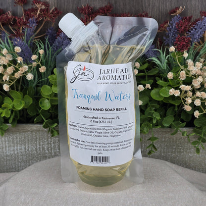A 16 fl oz (473 mL) clear pouch of Jarhead Aromatics Tranquil Waters Foaming Hand Soap sits on a wooden surface, featuring a label and colorful flowers in the background. The refill is made with organic oils.
