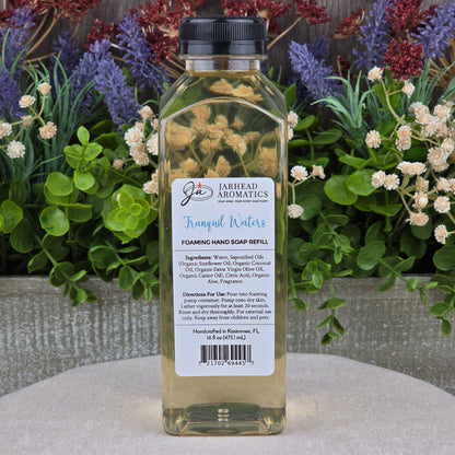 A clear rectangular bottle of Jarhead Aromatics Tranquil Waters Foaming Hand Soap sits on a round surface, with colorful artificial flowers and greenery in the background. The label features ingredients and usage directions for this gentle, plant-based soap.