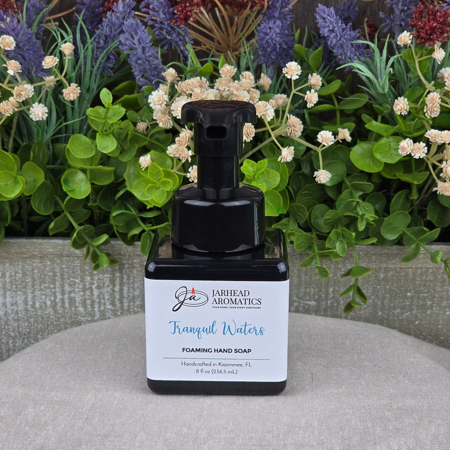 A bottle of Jarhead Aromatics Tranquil Waters Foaming Hand Soap rests on a tan surface, surrounded by green leaves, purple flowers, and white blossoms. The label features its handcrafted origin in Kissimmee, FL.