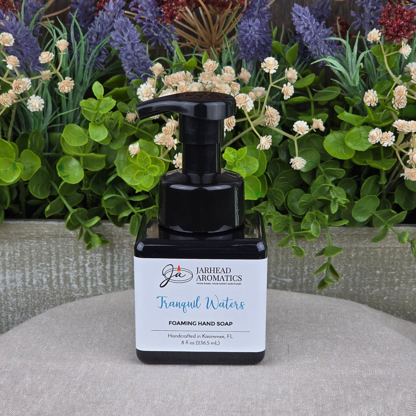 A bottle of Tranquil Waters Foaming Hand Soap by Jarhead Aromatics rests on wood, framed by green leaves and decorative purple and white flowers.