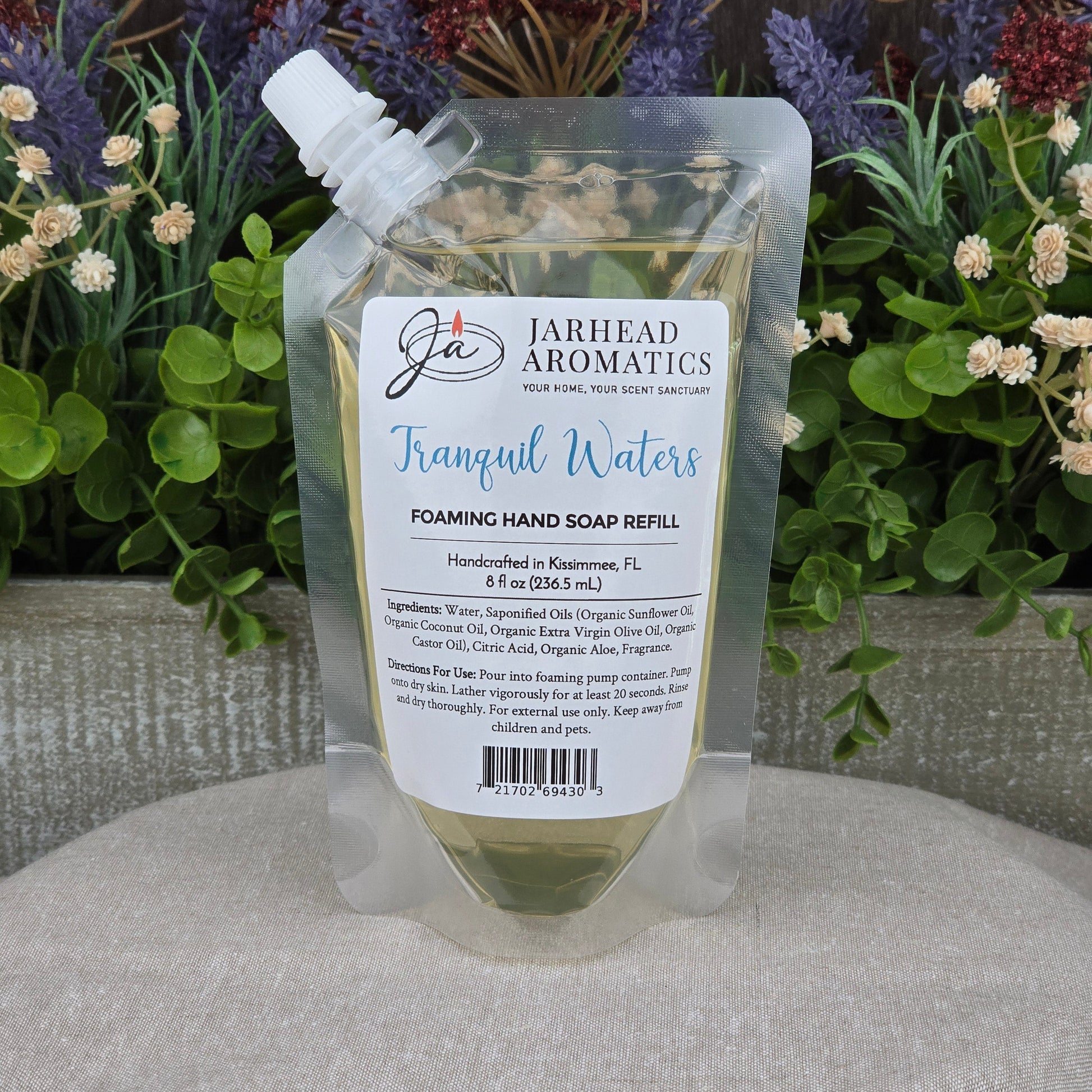 A clear pouch of Tranquil Waters Foaming Hand Soap by Jarhead Aromatics sits on a table, filled with pale liquid cleanser and set against colorful flowers and greenery.
