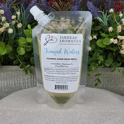 A clear pouch of Tranquil Waters Foaming Hand Soap by Jarhead Aromatics sits on a table, filled with pale liquid cleanser and set against colorful flowers and greenery.
