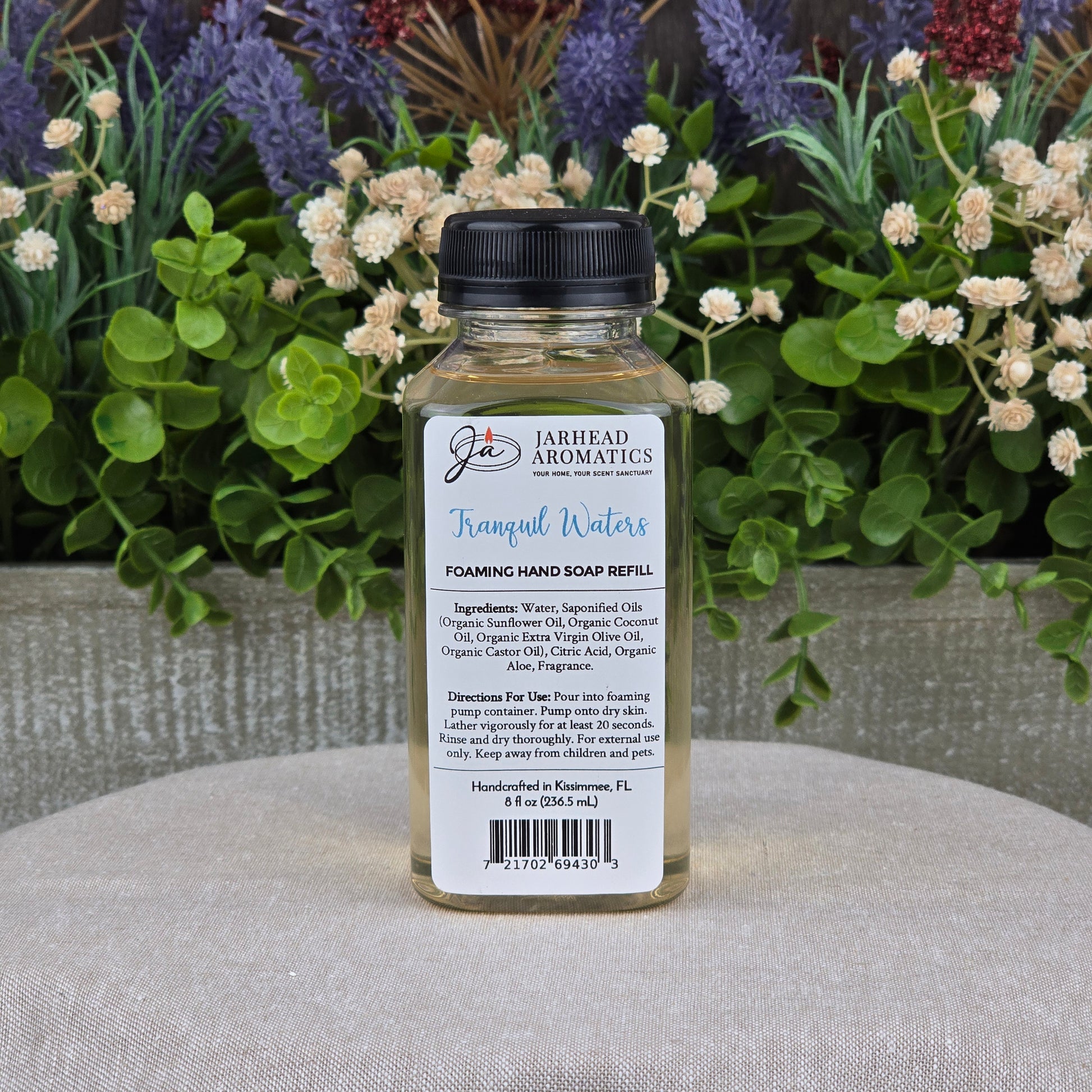 A clear bottle of Jarhead Aromatics Tranquil Waters Foaming Hand Soap sits on a gray surface, surrounded by green foliage and white and purple flowers. The label displays its gentle, plant-based ingredients and usage instructions.