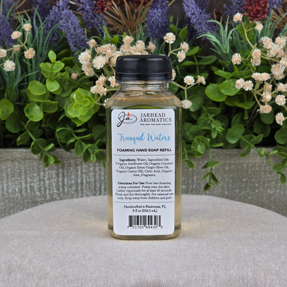 A clear bottle of Jarhead Aromatics Tranquil Waters Foaming Hand Soap sits on a gray surface, surrounded by green foliage and white and purple flowers. The label displays its gentle, plant-based ingredients and usage instructions.