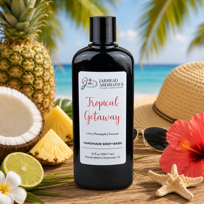 A bottle of Jarhead Aromatics Tropical Getaway Handmade Body Wash | Moisturizing Body Wash with Organic Aloe & Plant-Based Oils rests on the sand, surrounded by fresh pineapple, zesty lime, a sunhat, sunglasses, and a vibrant red flower. In the background, the beach and ocean stretch out beneath lush palm leaves—a captivating scene that perfectly highlights this luxurious organic body wash.