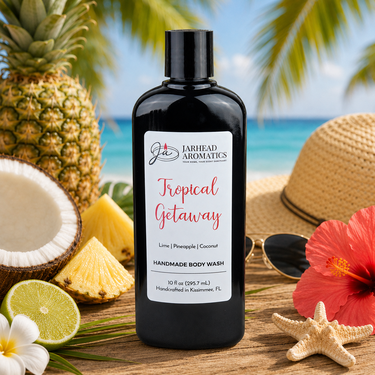 A bottle of Jarhead Aromatics Tropical Getaway Handmade Body Wash | Moisturizing Body Wash with Organic Aloe & Plant-Based Oils rests on sand amid tropical fruits, a hat, sunglasses, hibiscus flower, and palm trees beside the ocean.