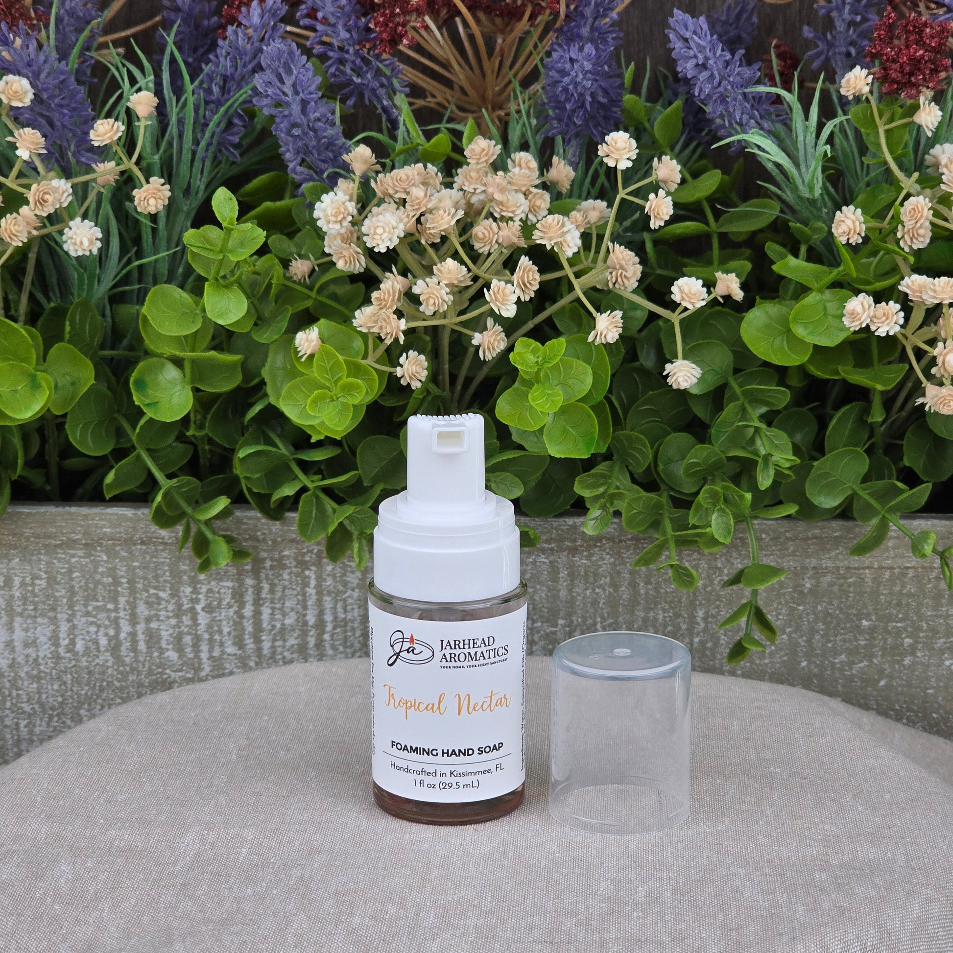 A bottle of Jarhead Aromatics Tropical Nectar Foaming Hand Soap, made with organic oils, stands on a beige surface with its clear cap beside it, in front of a lush arrangement of green leaves and purple flowers.