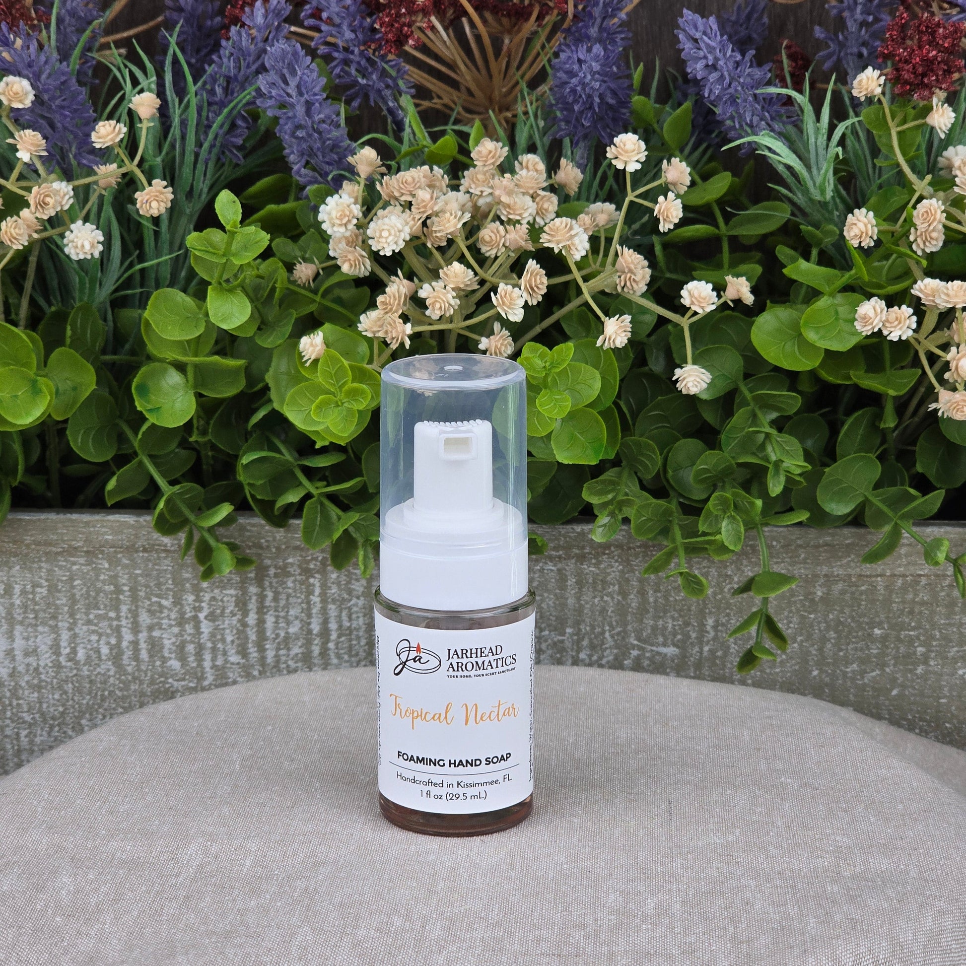 A bottle of Jarhead Aromatics Tropical Nectar Foaming Hand Soap sits on a gray cloth, surrounded by green leaves and purple and cream artificial flowers in the background.