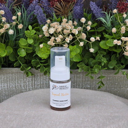 A bottle of Jarhead Aromatics Tropical Nectar Foaming Hand Soap sits on a gray cloth, surrounded by green leaves and purple and cream artificial flowers in the background.