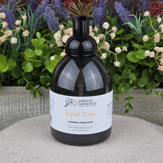 A bottle of Jarhead Aromatics Tropical Nectar Foaming Hand Soap rests on a gray surface, with leafy plants and purple and white flowers in the background.