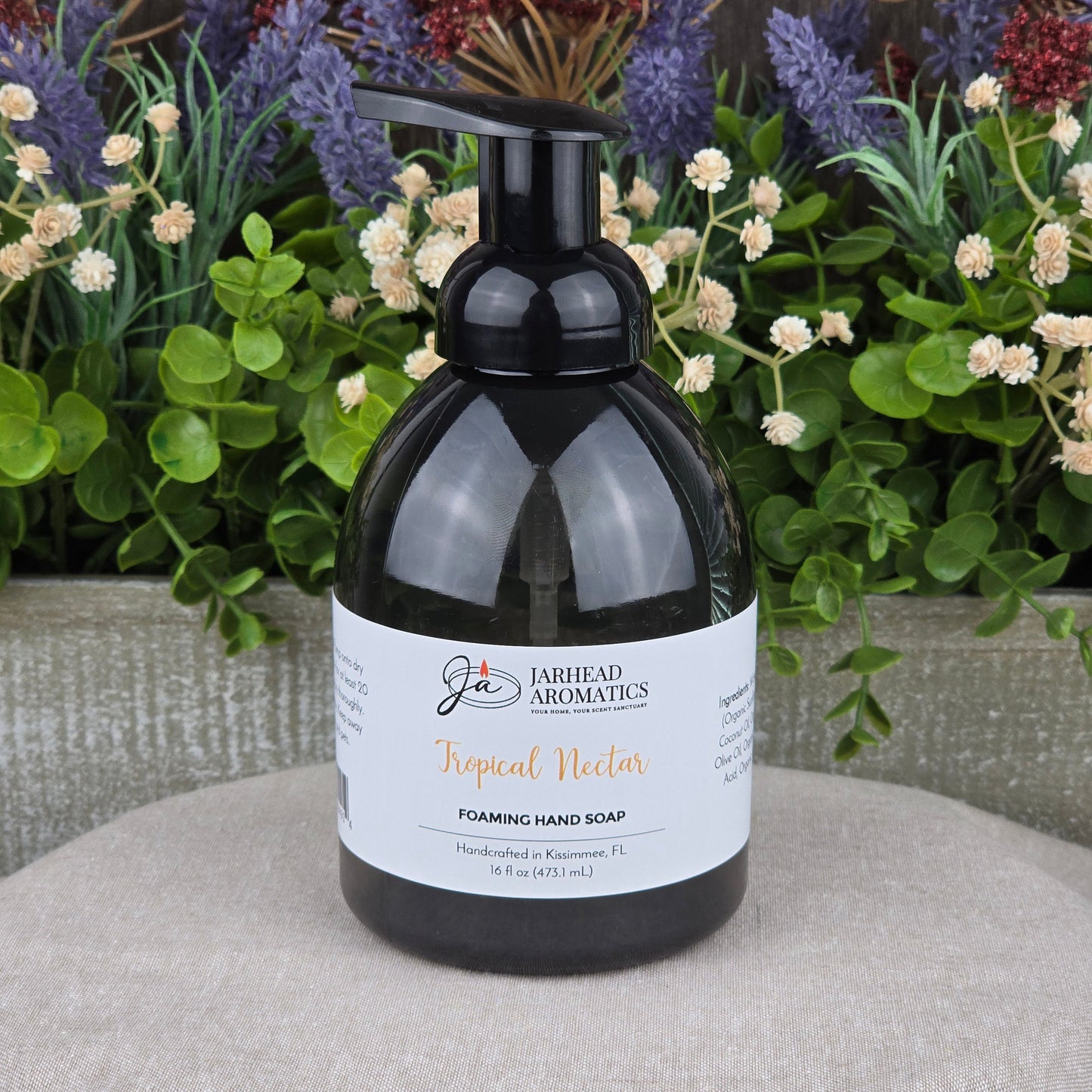 A black pump bottle of Jarhead Aromatics Tropical Nectar Foaming Hand Soap rests on a textured gray surface, surrounded by green leaves and colorful purple and red flowers in the background.