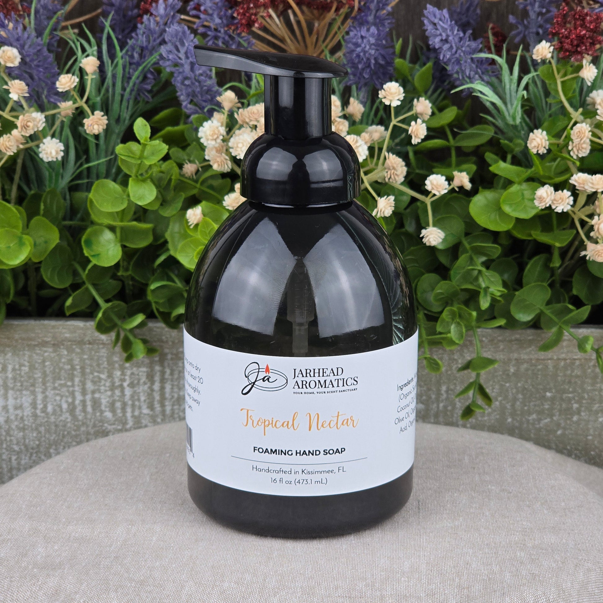 A black pump bottle of Jarhead Aromatics Tropical Nectar Foaming Hand Soap rests on a textured gray surface, surrounded by green leaves and colorful purple and red flowers in the background.