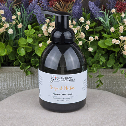 A black pump bottle of Jarhead Aromatics Tropical Nectar Foaming Hand Soap rests on a textured gray surface, surrounded by green leaves and colorful purple and red flowers in the background.