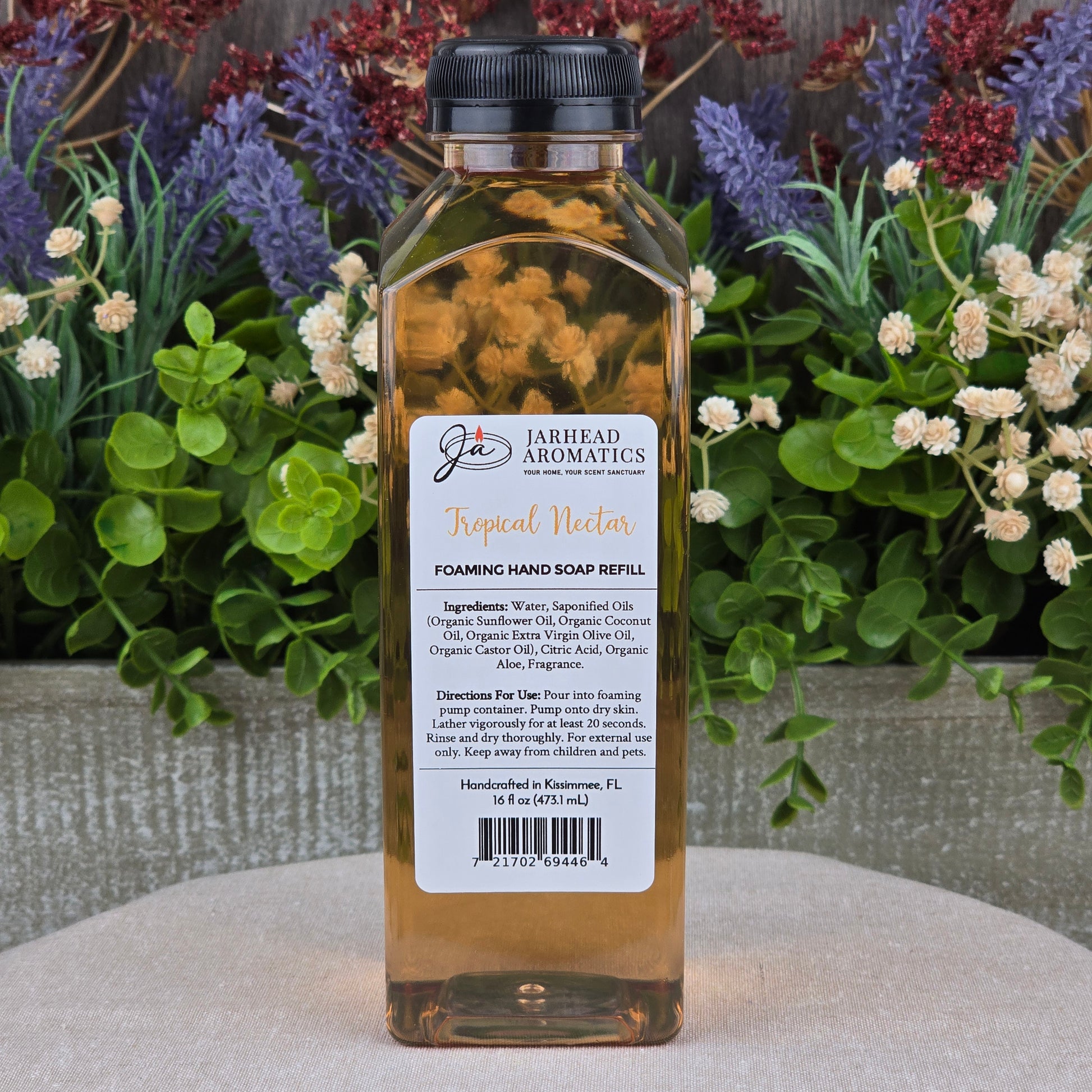 A clear rectangular bottle of Jarhead Aromatics Tropical Nectar Foaming Hand Soap rests on a stone surface amid vibrant flowers and greenery.