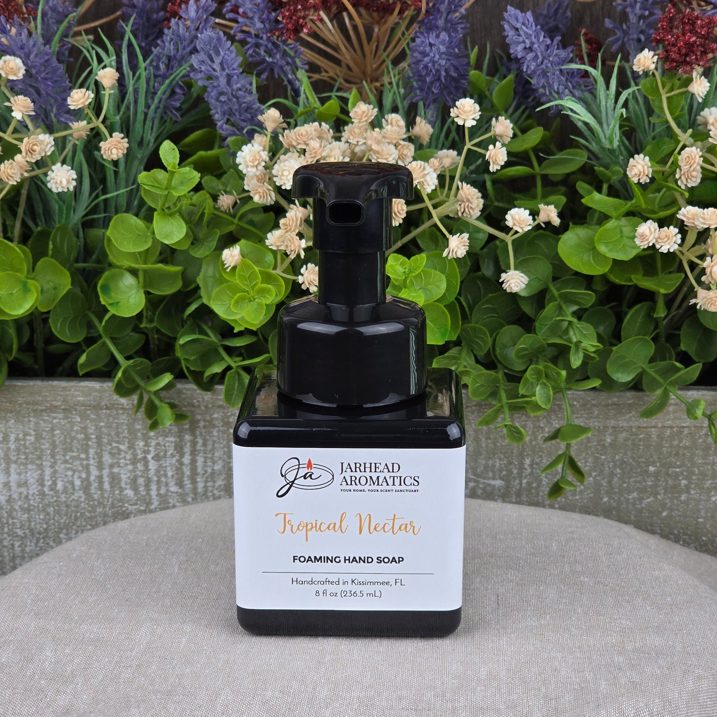 A black pump bottle of Jarhead Aromatics Tropical Nectar Foaming Hand Soap rests on a beige surface, surrounded by green leaves and bright artificial flowers. This plant-based cleanser is made with organic oils.