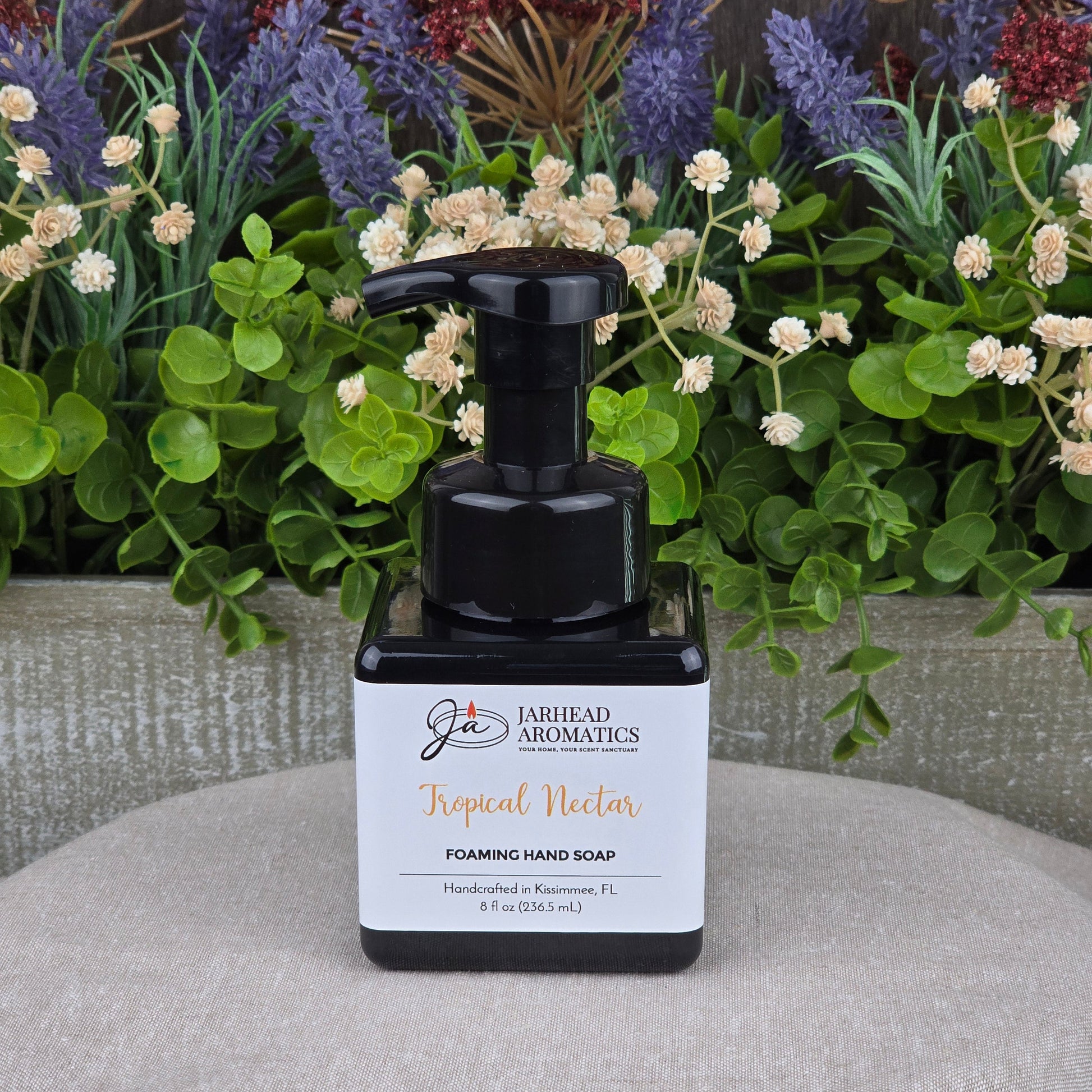 A black pump bottle of Jarhead Aromatics Tropical Nectar Foaming Hand Soap sits on a beige surface, backed by green and purple artificial plants and wood. This plant-based cleanser uses organic oils for gentle handwashing.