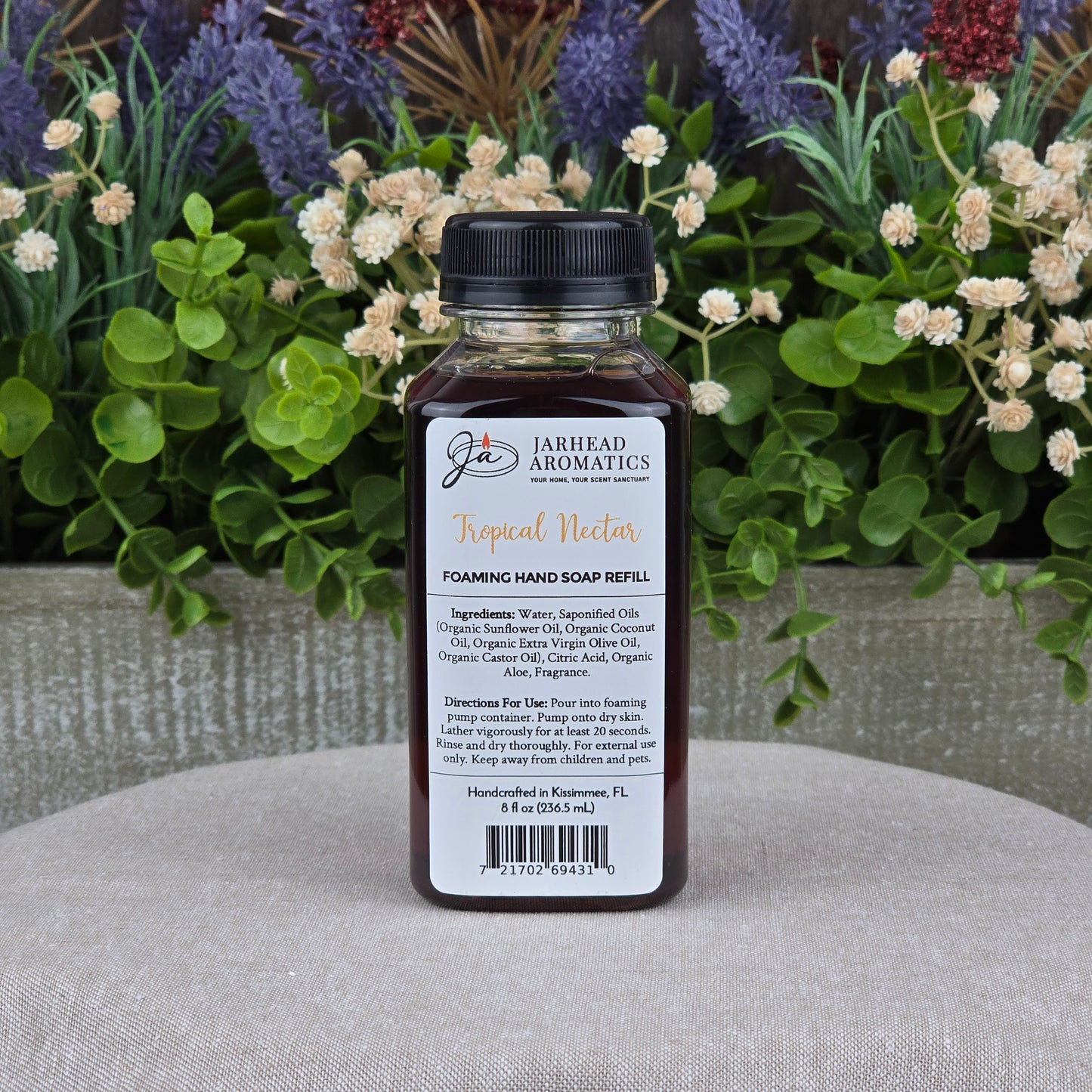 A bottle of Jarhead Aromatics Tropical Nectar Foaming Hand Soap, made with plant-based cleansers and organic oils, is shown on a textured surface with green foliage and purple flowers in the background.