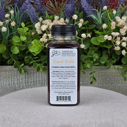 A bottle of Jarhead Aromatics Tropical Nectar Foaming Hand Soap, made with plant-based cleansers and organic oils, is shown on a textured surface with green foliage and purple flowers in the background.