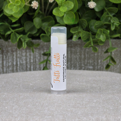 A tube of Jarhead Aromatics Tallow Handmade Natural Lip Balm in assorted flavors stands upright on a gray surface, with green leafy plants in the background.