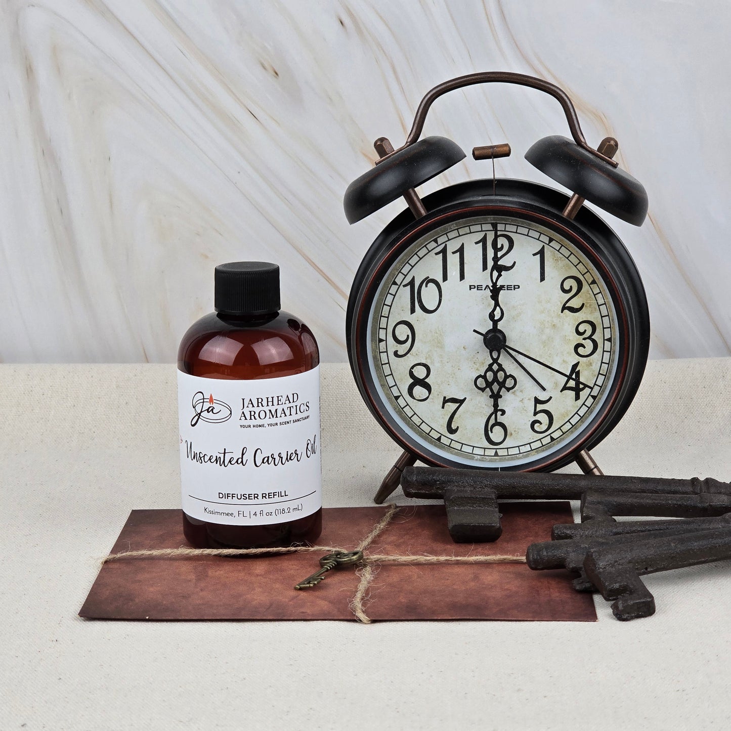 A vintage alarm clock, a brown bottle of Jarhead Aromatics Unscented Carrier Oil Diffuser Refill, old-fashioned keys, and a leather-bound book rest on a light surface with a marble background.