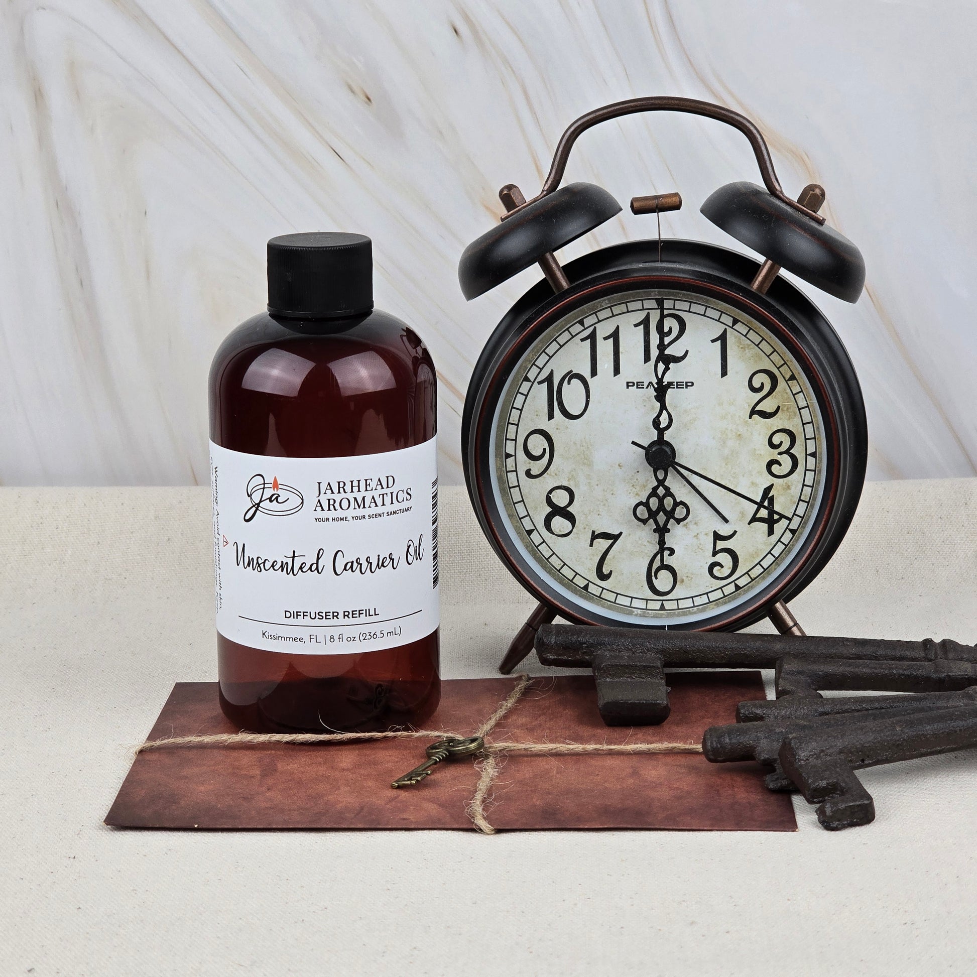 A bottle of Jarhead Aromatics Unscented Carrier Oil Diffuser Refill | Base Oil for Reed & Cold Air Diffusers is placed beside a vintage black alarm clock, three antique keys, and a brown envelope on a light marble-background surface.