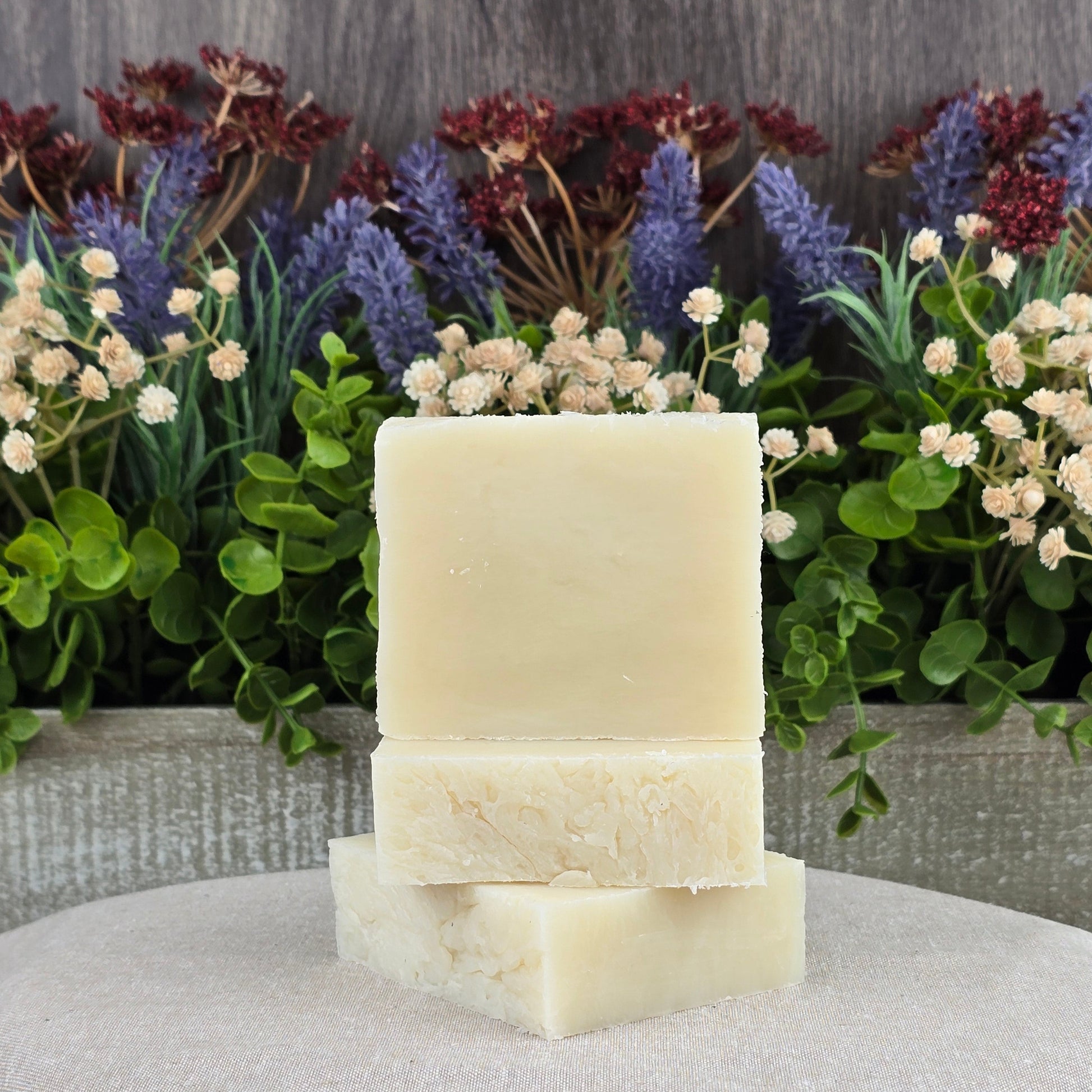 Three bars of Jarhead Aromatics Unscented Natural Handmade Soap - All Natural, ideal for sensitive skin, are stacked on neutral fabric before green leaves and flowers with a wooden background. Crafted with plant-based ingredients.