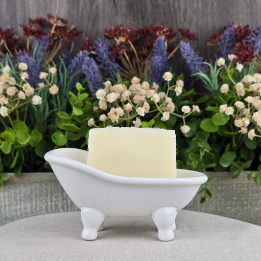 A white bar of Jarhead Aromatics Unscented Natural Handmade Soap - All Natural, ideal for sensitive skin, sits in a small white clawfoot bathtub-shaped dish with assorted artificial flowers and greenery behind it.