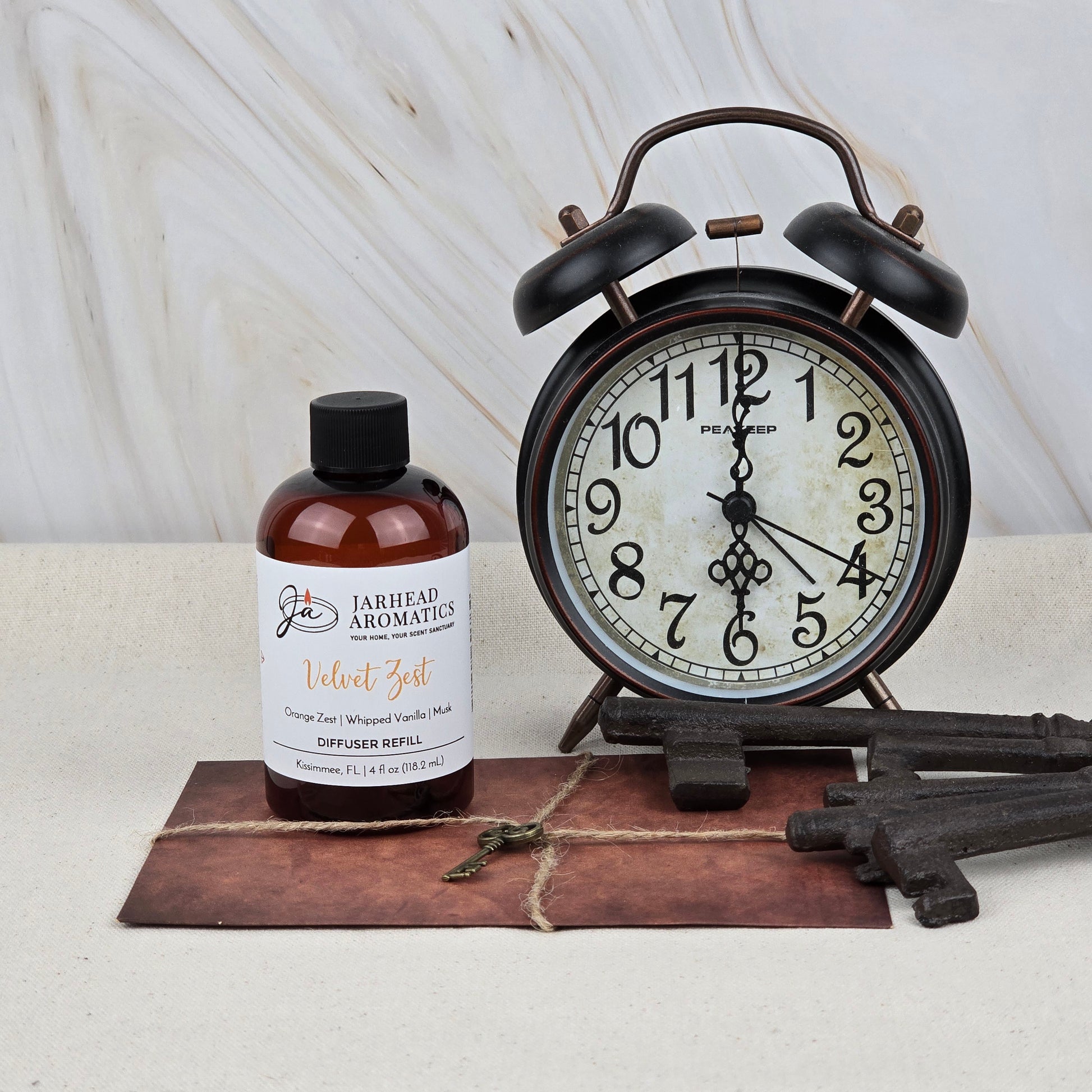 A brown bottle of Jarhead Aromatics Velvet Zest Diffuser Refill, a citrus, vanilla & brown sugar fragrance oil, is displayed beside a vintage black alarm clock, three antique keys, and an old book with a small key on top.