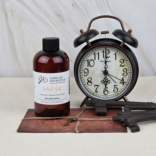 A brown bottle of Jarhead Aromatics Velvet Zest Diffuser Refill | Citrus, Vanilla & Brown Sugar Home Fragrance Oil sits beside a vintage black alarm clock, old metal keys, and a brown notepad on a neutral surface with a light background.