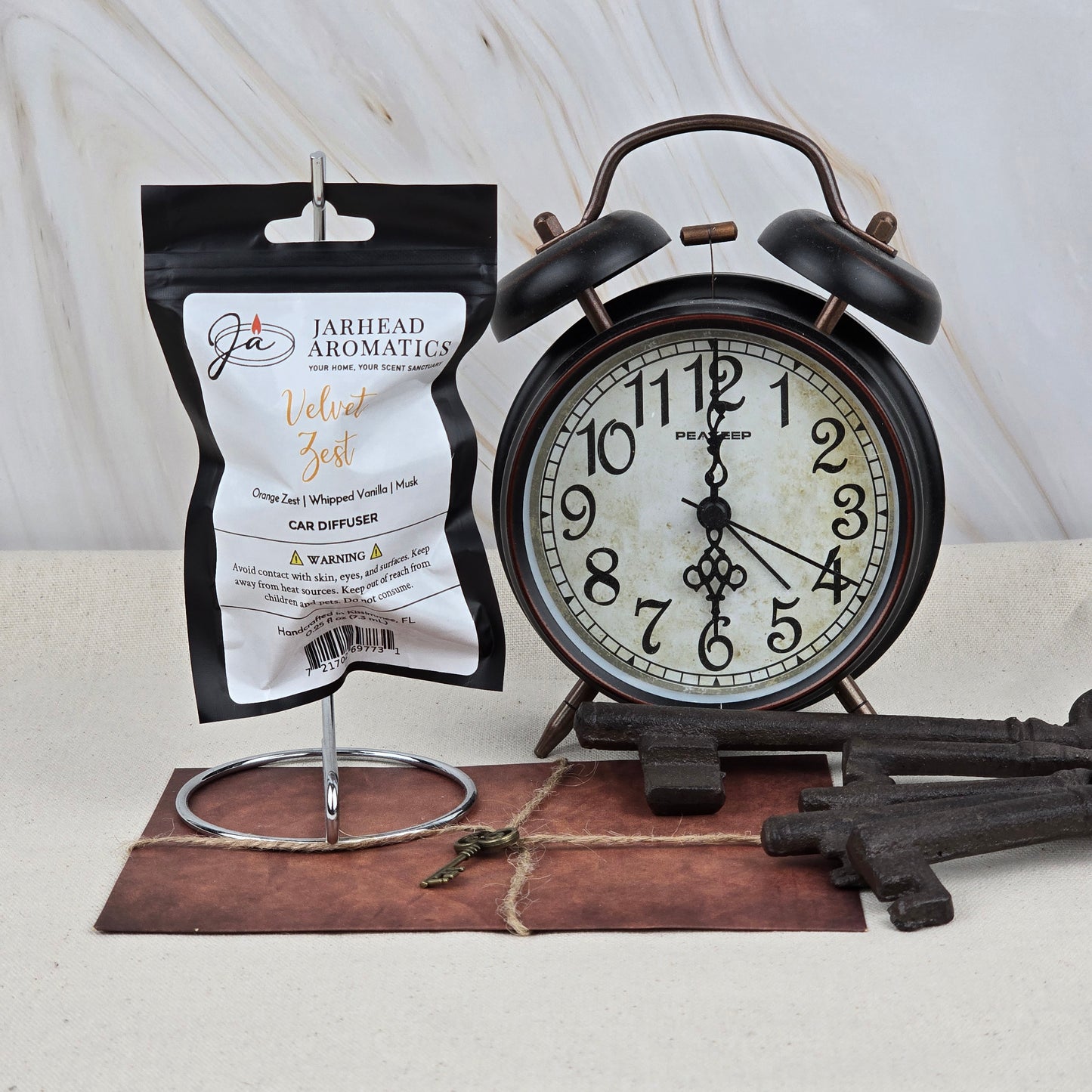 A black vintage alarm clock rests on a brown mat beside Jarhead Aromatics’ Velvet Zest Car Diffuser—Citrus Vanilla & Brown Sugar—renowned for its long-lasting scent, with metal keys nearby on a neutral background.