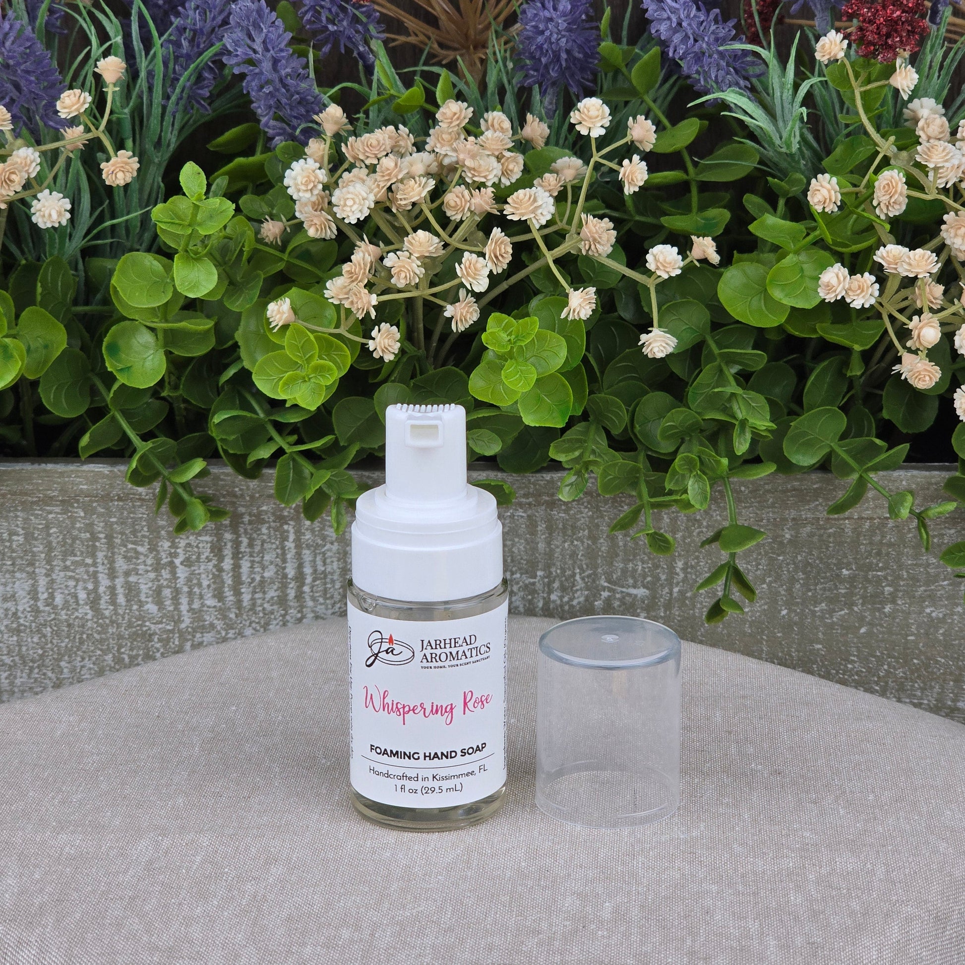 A bottle of Jarhead Aromatics Whispering Rose Foaming Hand Soap, with its clear cap off, sits on a beige surface before artificial greenery and flowers. Plant-based oils provide a gentle, coastal scent.