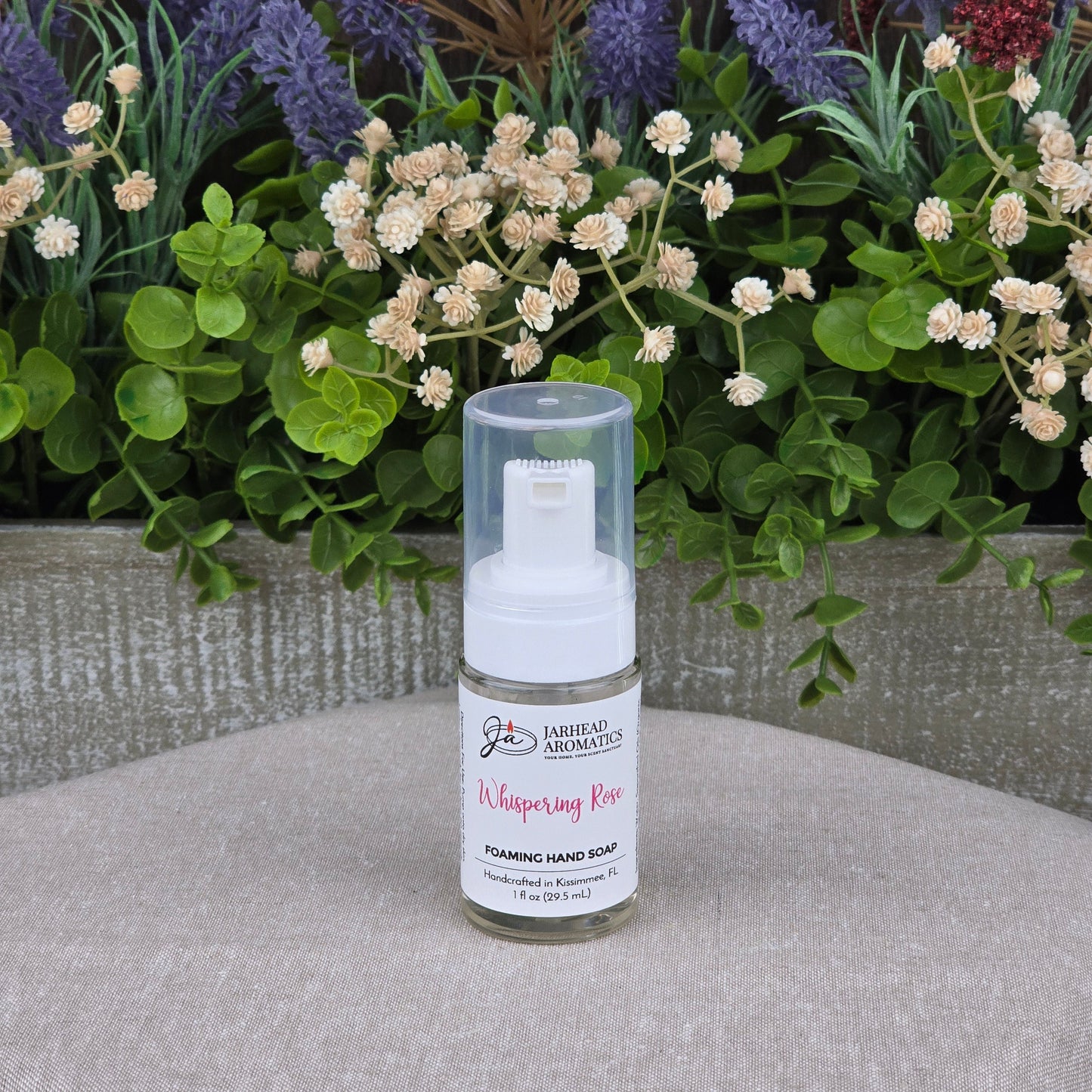 A bottle of Jarhead Aromatics Whispering Rose Foaming Hand Soap, made with plant-based oils, sits on a beige surface with green leaves and purple, white, and red artificial flowers in the background.