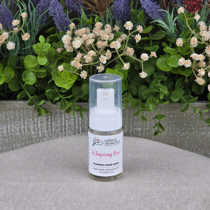A bottle of Jarhead Aromatics Whispering Rose Foaming Hand Soap, made with plant-based oils, sits on a beige surface with green leaves and purple, white, and red artificial flowers in the background.