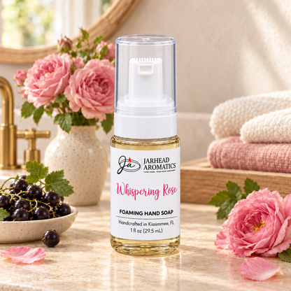 A bottle of Whispering Rose Handmade Foaming Hand Soap | Black Currant, Turkish Rose & White Musk by Jarhead Aromatics surrounded by fresh black currants and pink roses on a vanity counter.