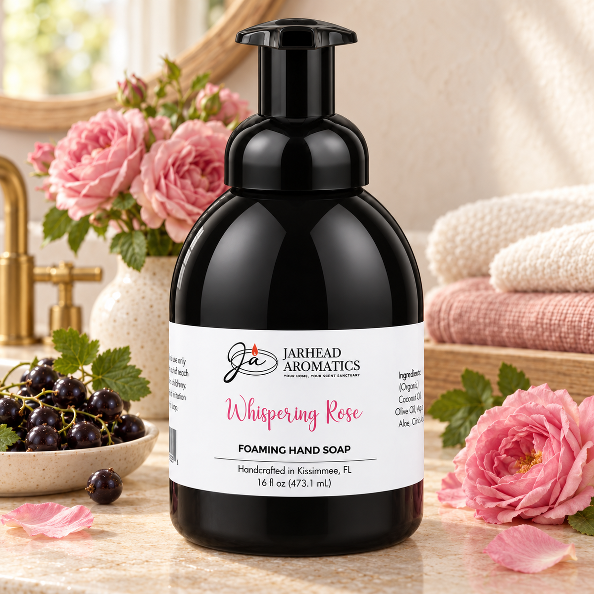 A round bottle of Jarhead Aromatics Whispering Rose Handmade Foaming Hand Soap, filled with black soap and surrounded by black currants and pink rose blossoms.