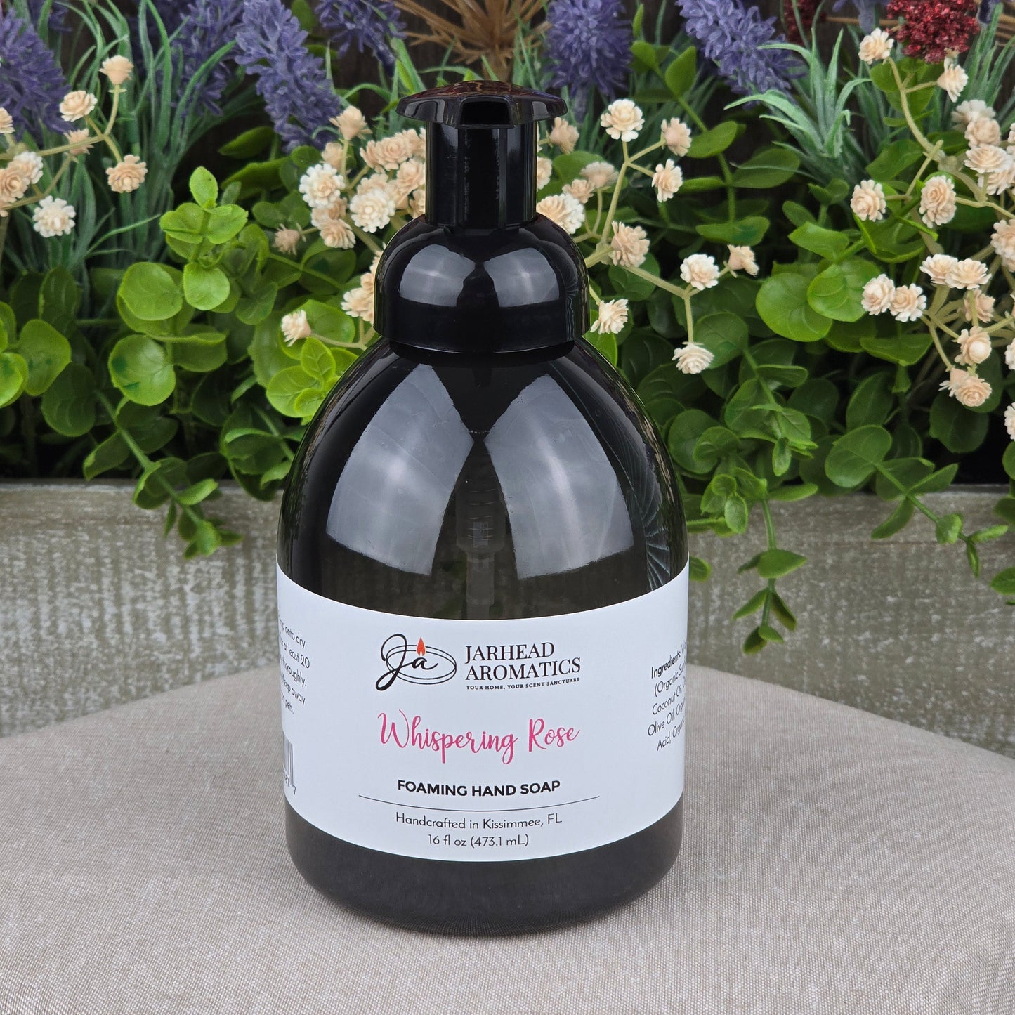 A black pump bottle of Jarhead Aromatics Whispering Rose Foaming Hand Soap rests on a round stone surface, with colorful artificial flowers and greenery in the background.