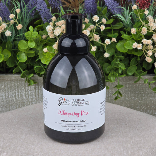 A black pump bottle of Jarhead Aromatics Whispering Rose Foaming Hand Soap rests on a round stone surface, with colorful artificial flowers and greenery in the background.