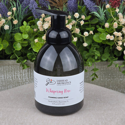 A bottle of Jarhead Aromatics Whispering Rose Foaming Hand Soap rests on a gray surface, with colorful flowers and greenery softly blurred in the background.