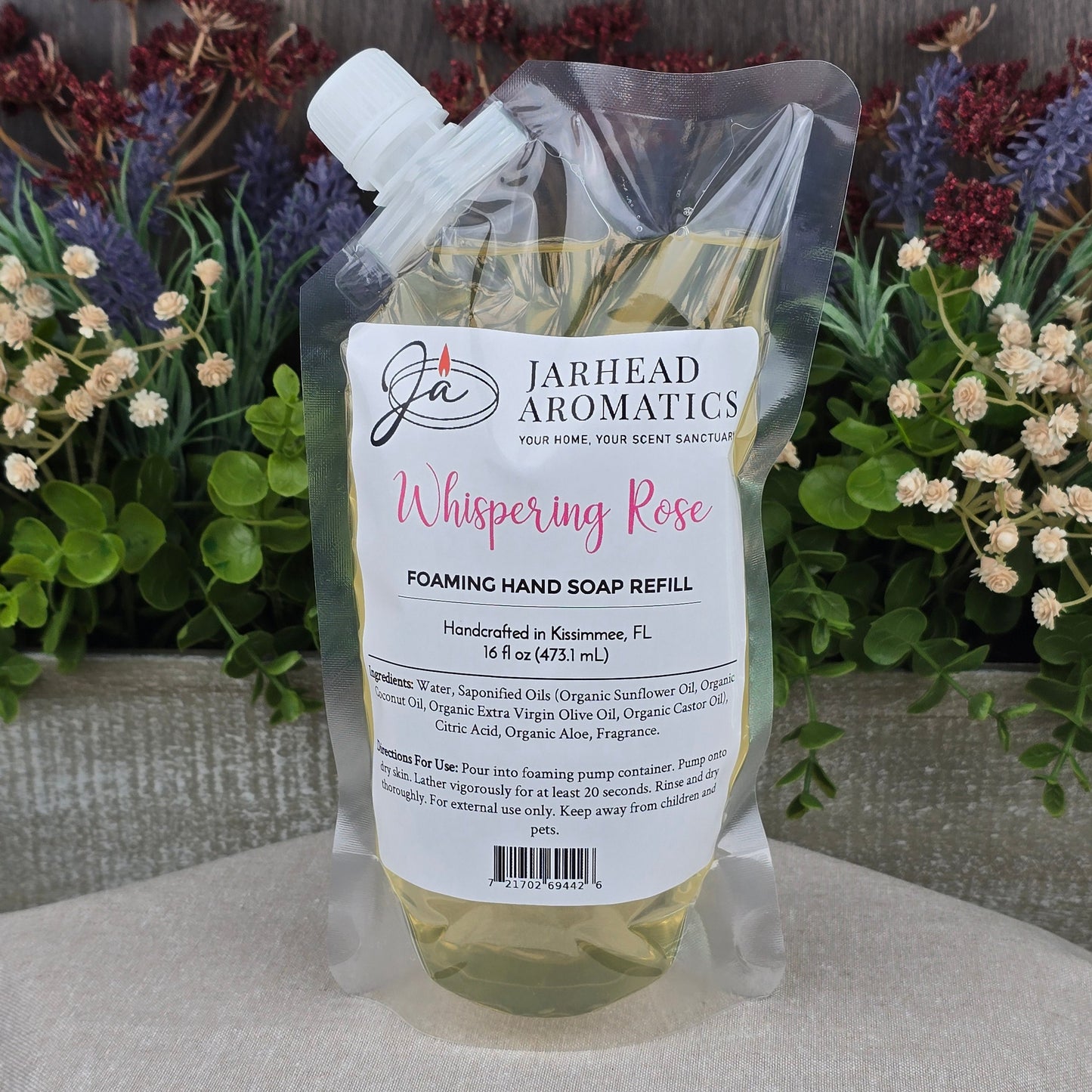 A clear pouch of Jarhead Aromatics Whispering Rose Foaming Hand Soap sits on a beige surface with green and purple faux flowers, a wooden backdrop, and boasts gentle plant-based oils.