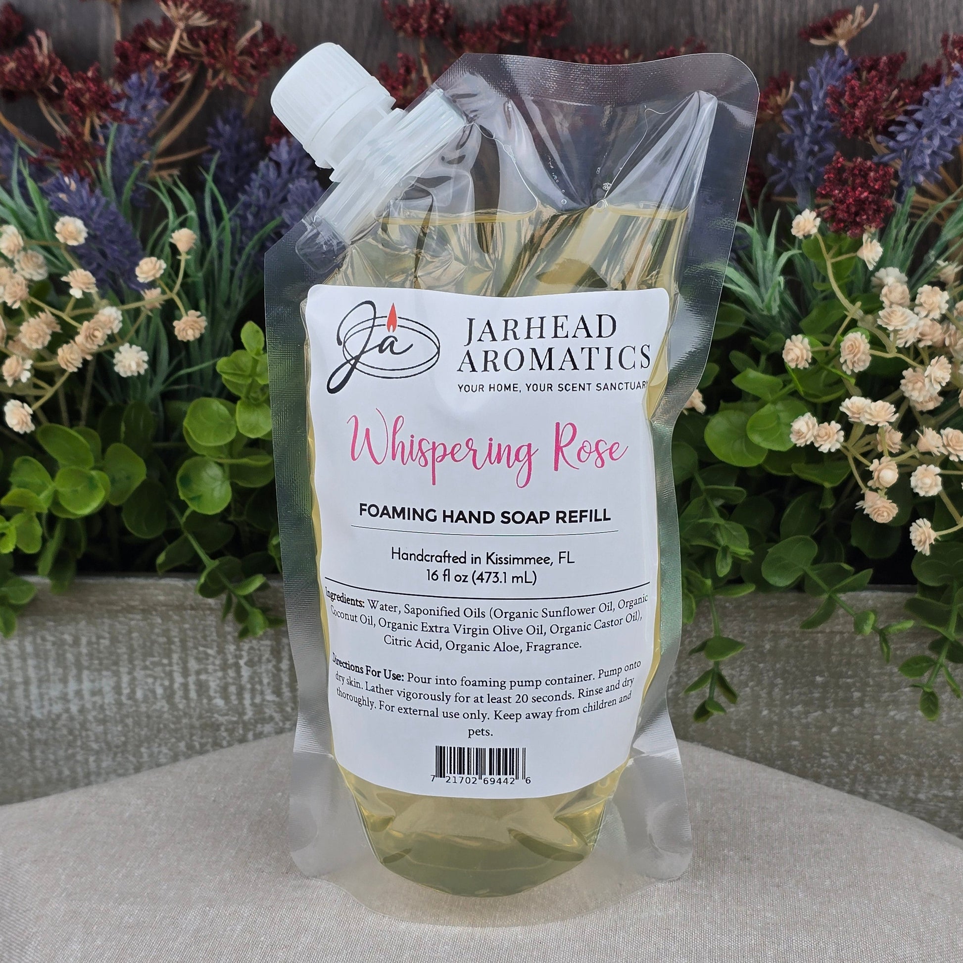 A clear pouch of Jarhead Aromatics Whispering Rose Foaming Hand Soap sits on a beige surface with green and purple faux flowers, a wooden backdrop, and boasts gentle plant-based oils.
